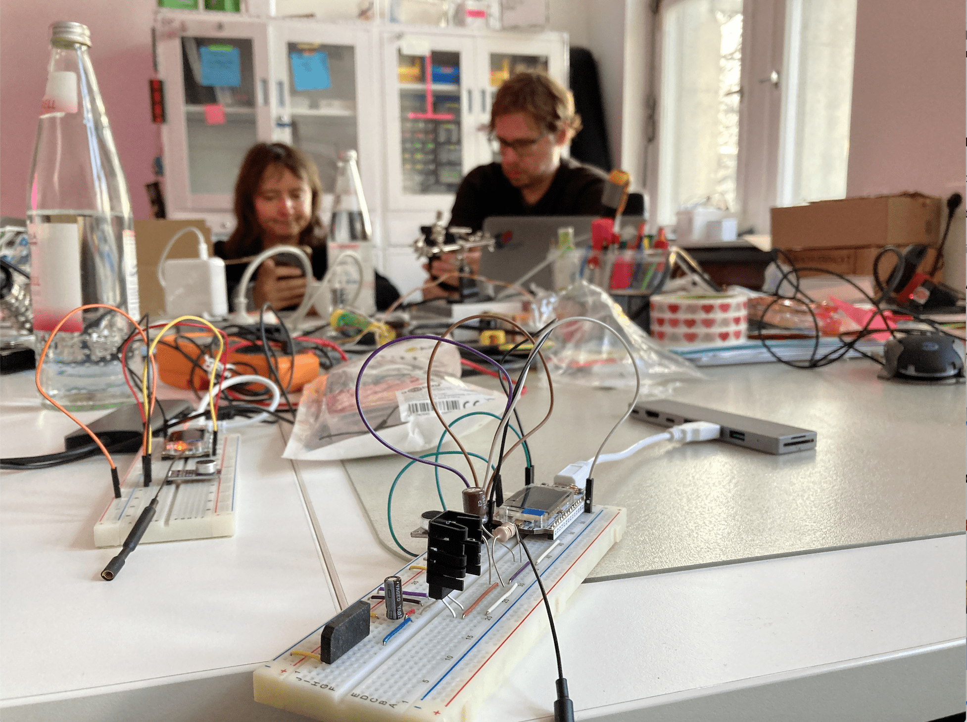 Boards, sensors, and cables are on top of a table while two people are working in the background