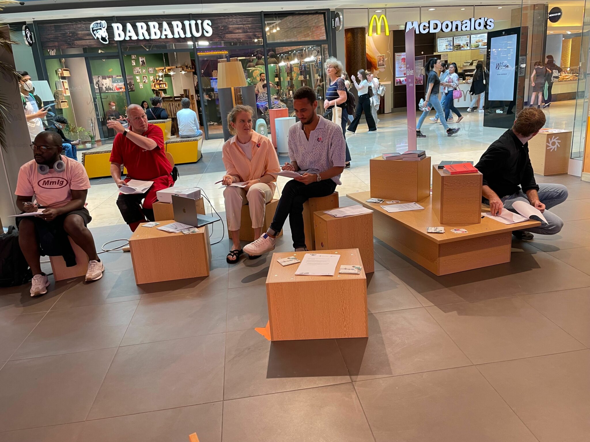 5 people sitting in the Mall, reading and talking to each other. There are stores and people walking by in the background.