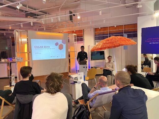 A group of people gathered and listening to a presentation. In the center are presentation slides, the speaker and a big orange parasol.