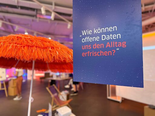 You see a close-up of sign saying "How can Open Data help to freshen up our every day live" - refering to the project "Erfrischungskarte" by ODIS Berlin. In the background you see a deck chair and a big orange parasol.