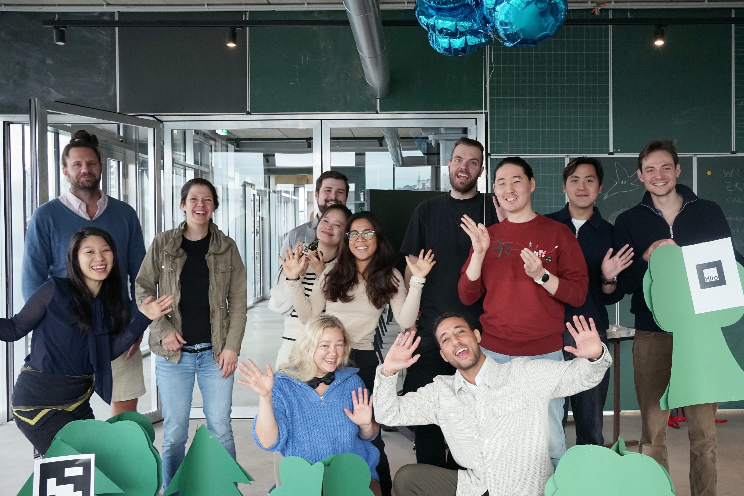 Gruppe von Teilnehmenden der Summer School 2025, die fröhlich in die Kamera lächeln und winken, aufgenommen bei der Abschlussveranstaltung im CityLAB Berlin.