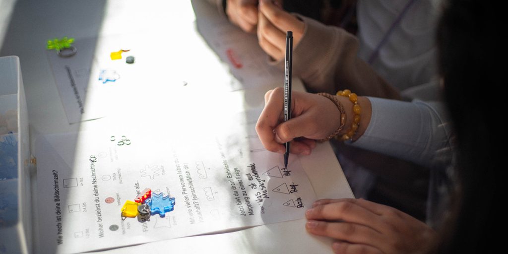 Workshop-Teilnehmerin beim Girlsday füllt mit einem schwarzen Stift einen Fragebogen aus, auf dem Tisch liegen bunte Kunststoffanhänger, kleine Metallringe und weiteres Bastelmaterial.