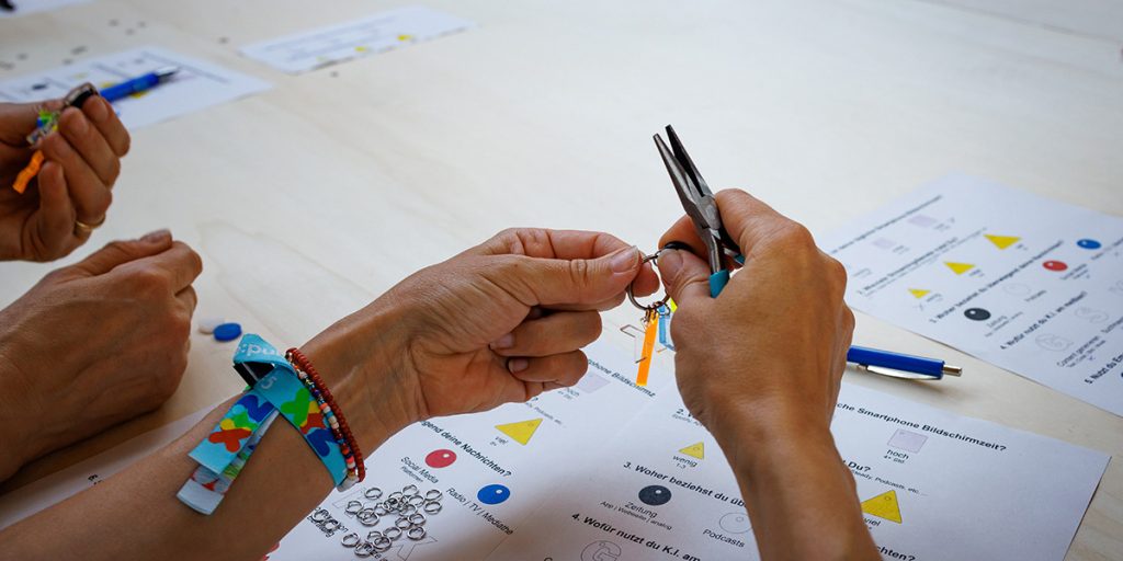 Gruppe von Workshop-Teilnehmenden steht an einem langen Tisch, tauschen sie aus und bastelt bunte Schlüsselanhänger auf der re:publica 2025.