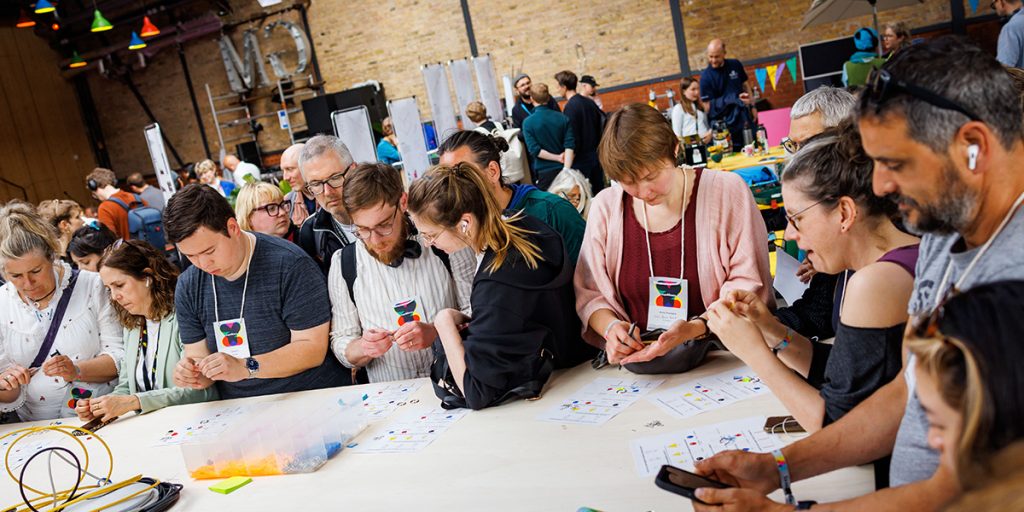 Gruppe von Workshop-Teilnehmenden steht an einem langen Tisch, tauschen sie aus und bastelt bunte Schlüsselanhänger auf der re:publica 2025.