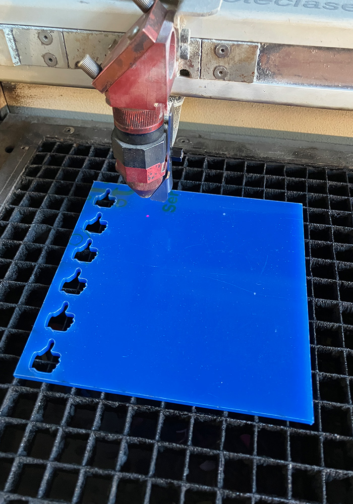 Nozzel eines Lasercutters, der kleine Daumen-Hoch-Formen aus einem blauen Acrylglasstück herausschneidet.